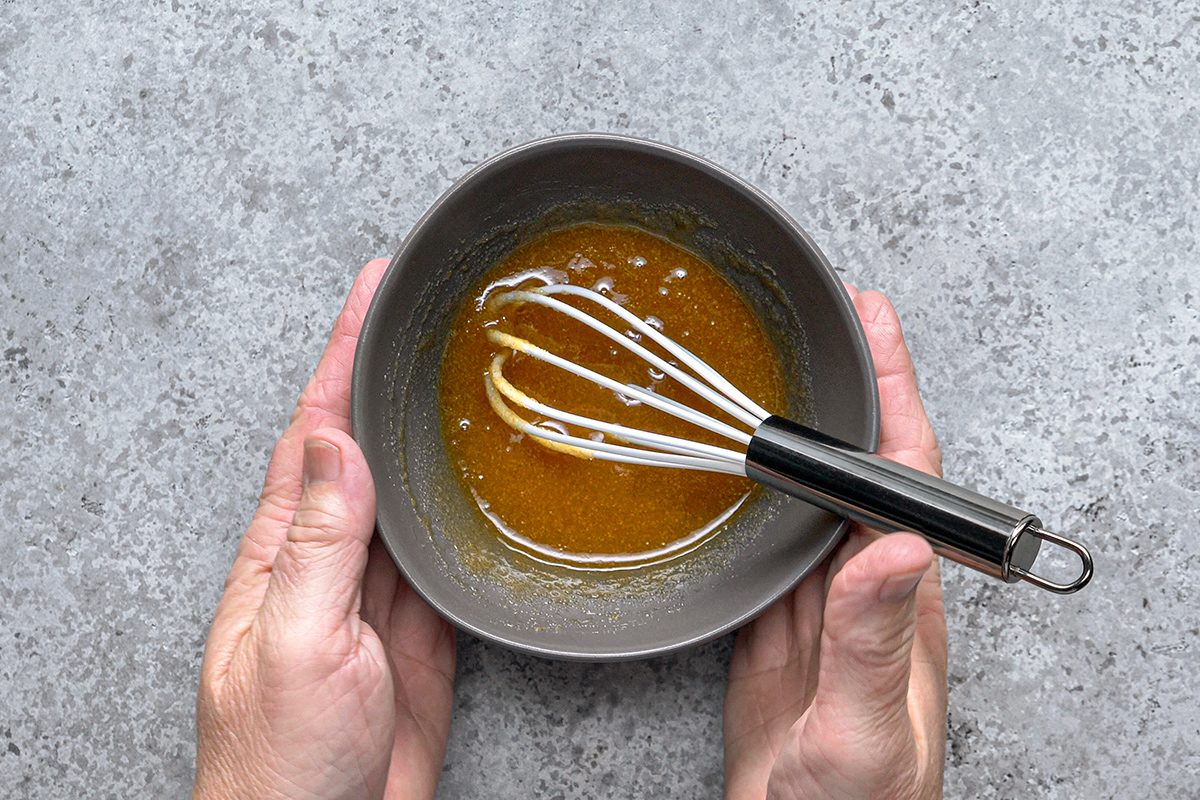 Two hands hold a gray bowl with a metal whisk mixing a golden-brown liquid on a gray textured surface.