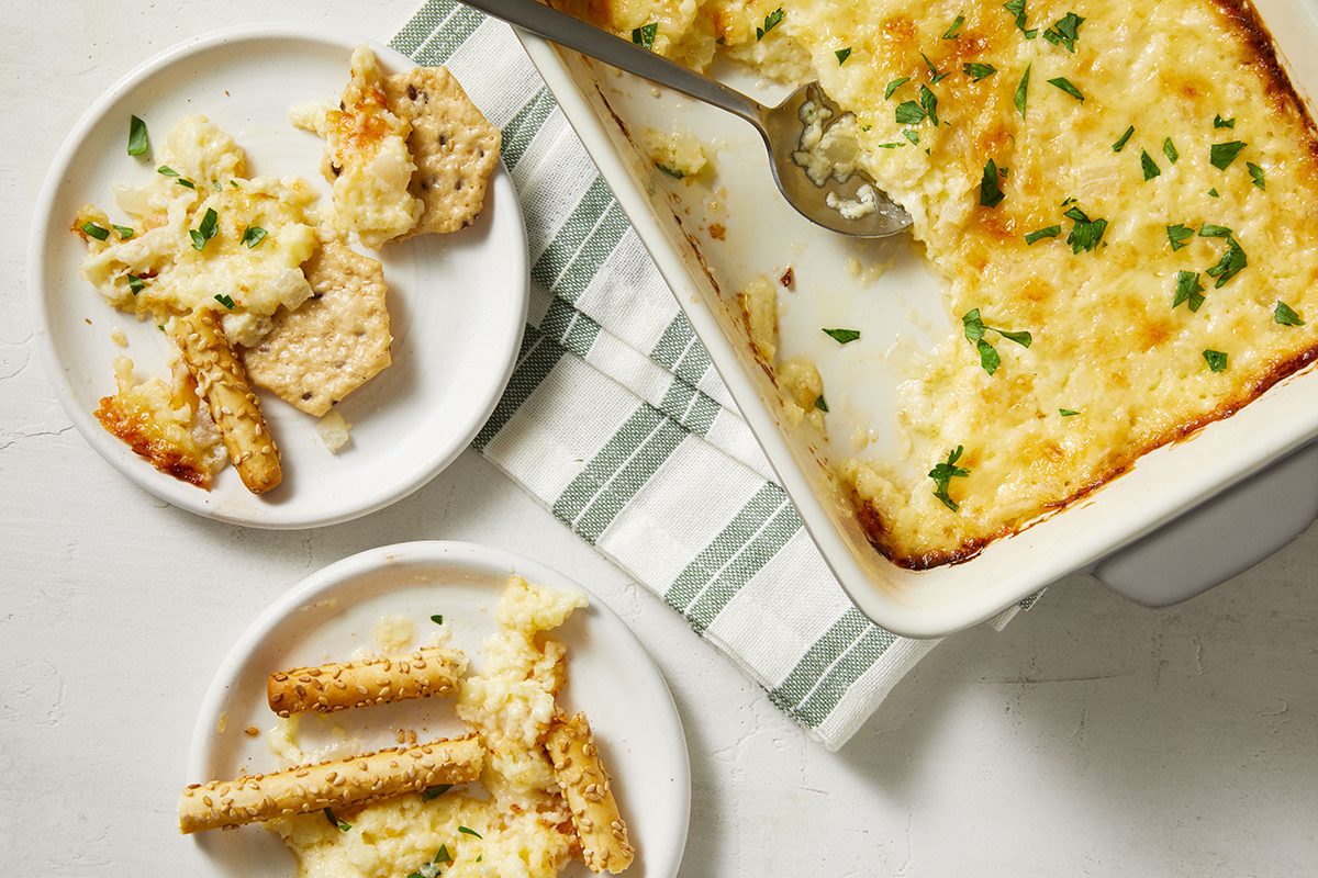 A baking dish of cheesy dip with a serving spoon, next to two small plates holding dip, crackers, and breadsticks. The dish and plates rest on a white and green striped towel on a white surface.