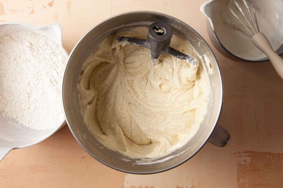 Overhead shot of a mixing bowl is filled with creamy cake batter and sits on a light brown surface and The left side shows a bowl of flour while the right displays a bowl with a whisk and whipped mixture