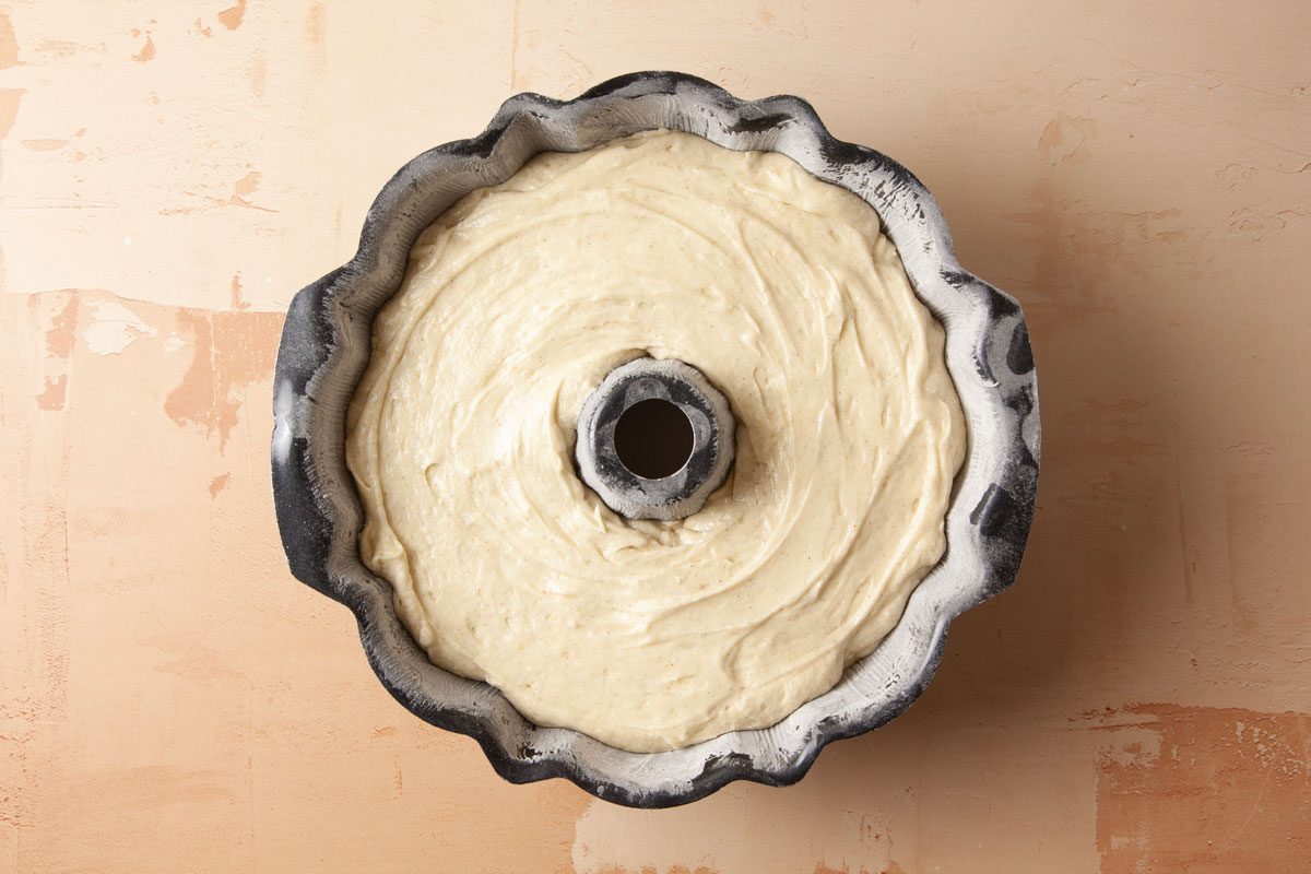 Overhead shot of a bundt cake pan holds smooth unbaked cake batter on a light tan textured surface and the pan is fluted and lightly coated with flour