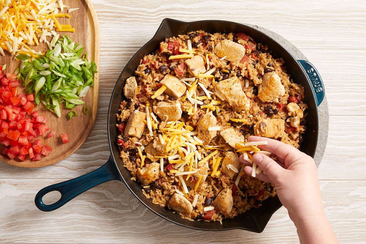 Overhead shot of shredded cheese and diced toppings being added to skillet by hand.