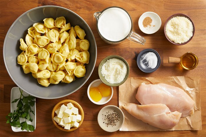 Overhead shot of ingredients laid out on a wooden surface including cheese tortellini, chicken breasts, milk, butter, parmesan, garlic, and herbs.