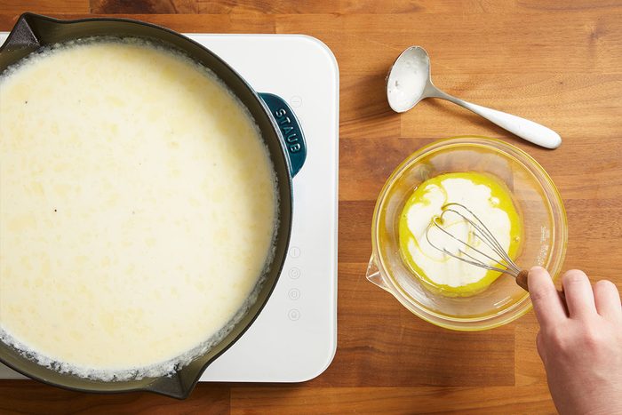 Overhead shot of butter being added to a simmering pan of Alfredo sauce with a spoon nearby.