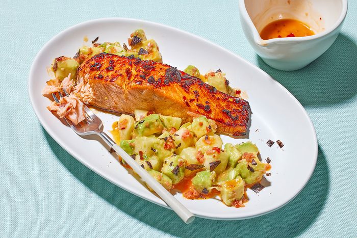 3/4 angle view shot of chili crisp glazed salmon served with a chunky avocado and cucumber salad on an oval white plate, with a fork placed alongside and an empty sauce bowl in the background;