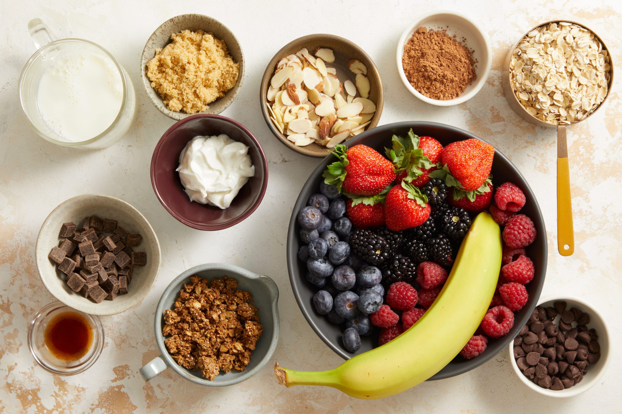 Overhead shot of various ingredients for chocolate overnight oats—banana, berries, nuts, cocoa powder, oats, yogurt, and milk—arranged in small bowls.
