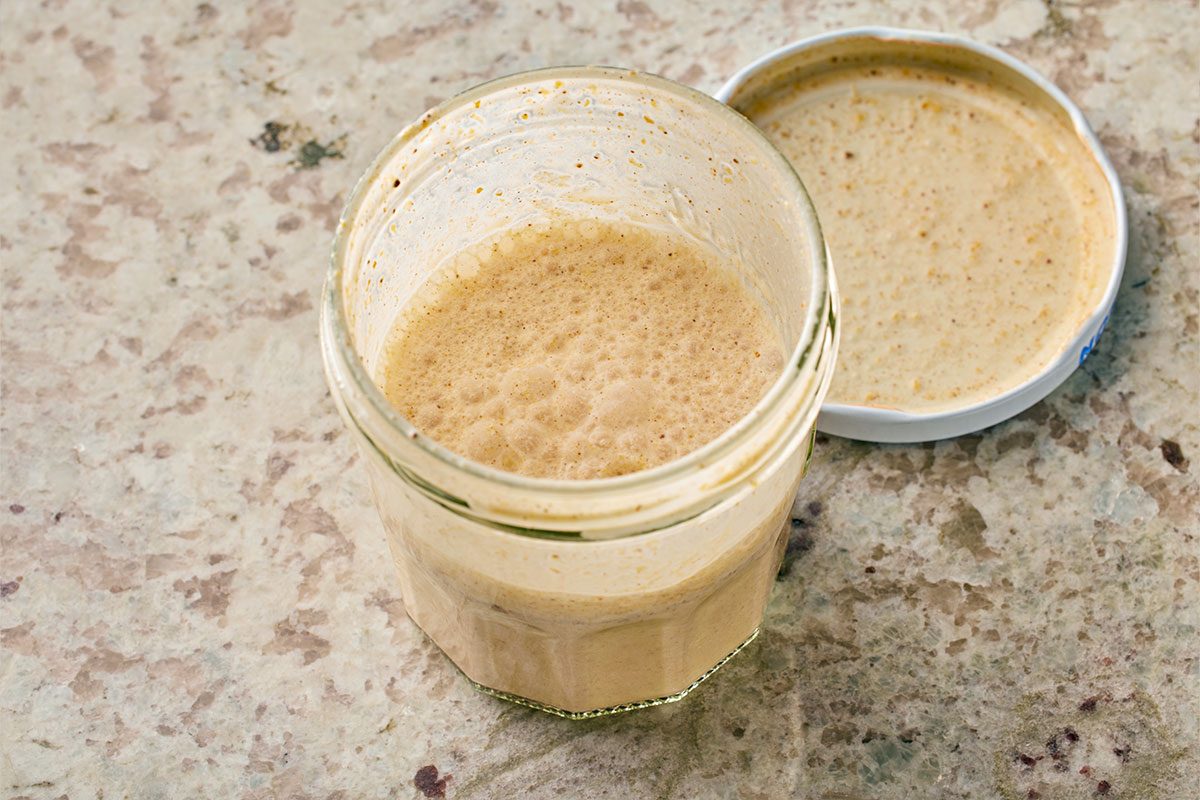 Overhead shot of an open glass jar filled with bubbly, light brown sourdough starter on a marbled countertop, with its matching lid beside it showing some starter residue;