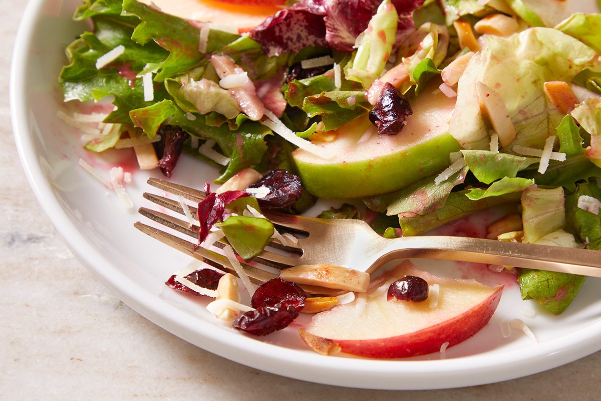 A close-up of a plate with a partially eaten salad, including lettuce, apple slices, dried cranberries, shredded cheese, and a fork resting on the side.