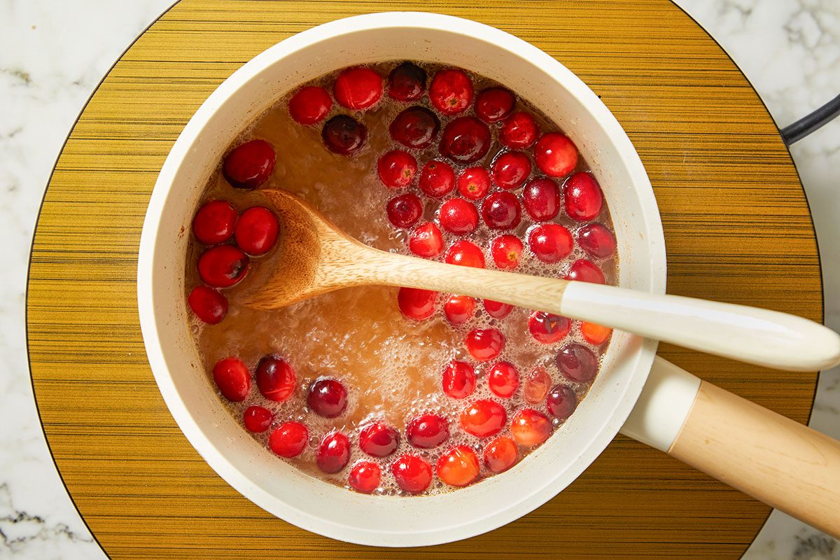 Overhead shot of cranberries simmering in a saucepan with sugar and water, a wooden spoon stirring the bubbling mixture.