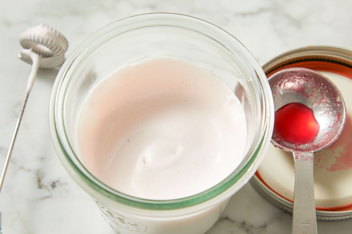 Overhead shot of a bowl of whipped cream tinted faintly pink from a spoonful of cranberry syrup, spoon resting inside.