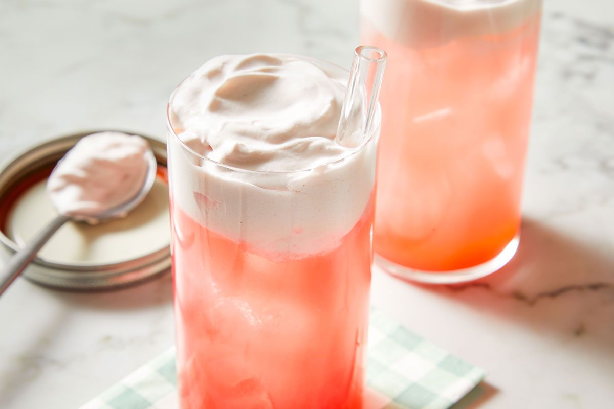 3/4 shot of two glasses of Cranberry Whipped Ginger Ale, creamy tops swirling into the pink soda, with a spoon coated in cranberry syrup nearby.