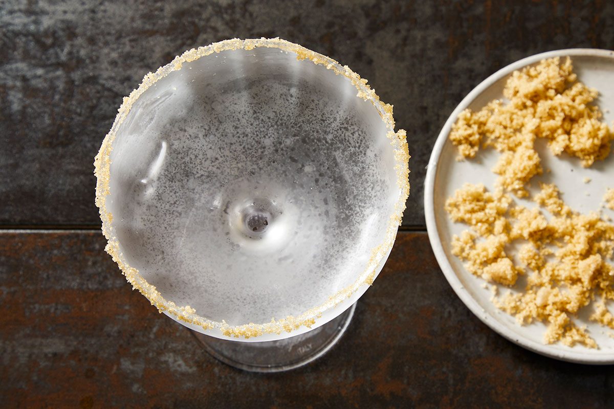 Overhead shot of a chilled cocktail glass with a sugared rim, set on a dark textured surface with sugar crystals nearby;