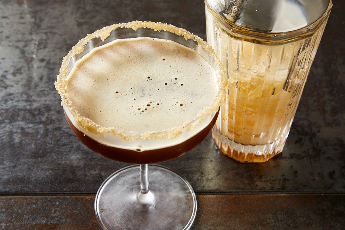 3/4 angle close-up shot of a creamy cocktail in a coupe glass with a sugared rim, set on a dark textured surface with a mixing glass beside it;