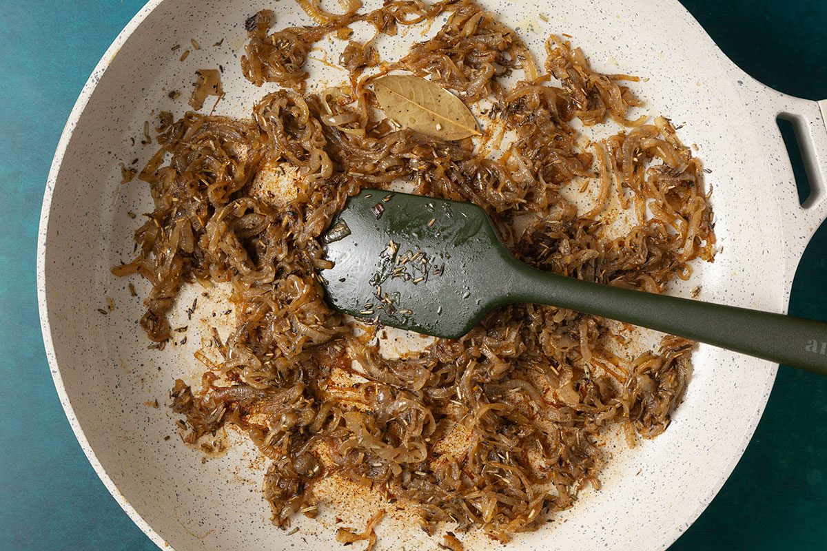 Overhead shot of caramelized onions cooking in a light-colored skillet, seasoned with spices and a bay leaf; A dark green spatula rests in the pan as the onions turn deep golden-brown against a teal surface;