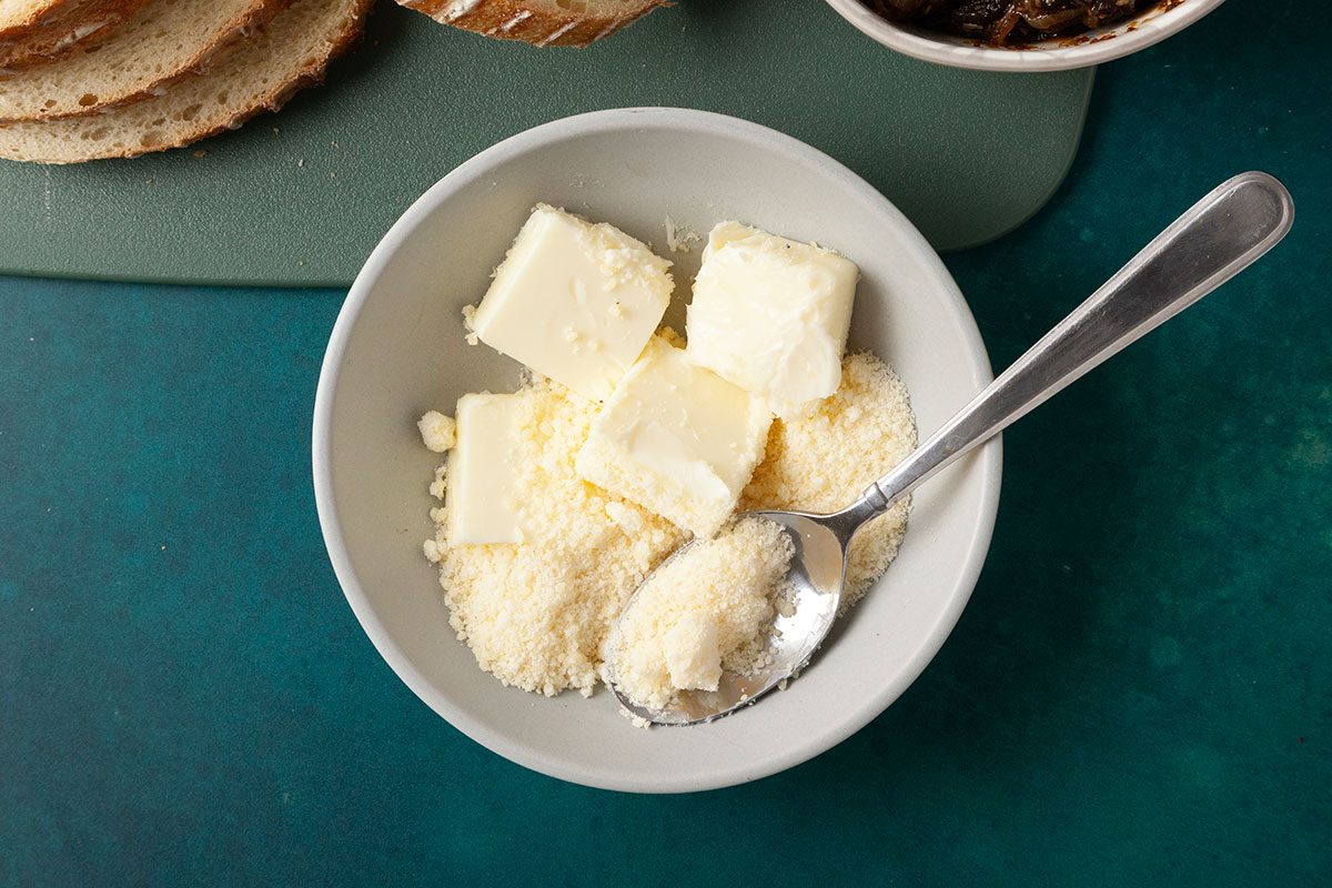 Overhead shot of a small gray bowl holding cubed butter and grated cheese with a metal spoon resting inside; Sliced bread sits on a cutting board nearby, with a bowl of caramelized onions in the background on a teal surface;