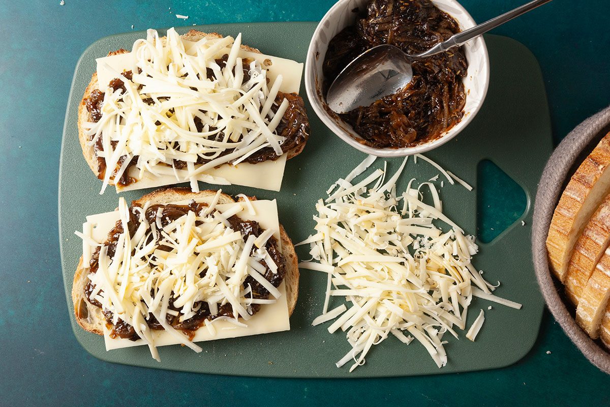 Overhead shot of Two slices of bread rest on a green cutting board topped with cheese and caramelized onions A bowl of onions and extra bread are close by