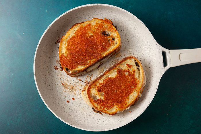 Overhead shot of two assembled sandwiches toasting in a light-colored skillet until golden brown; The bread is evenly crisped with a buttery surface, set against a teal background;