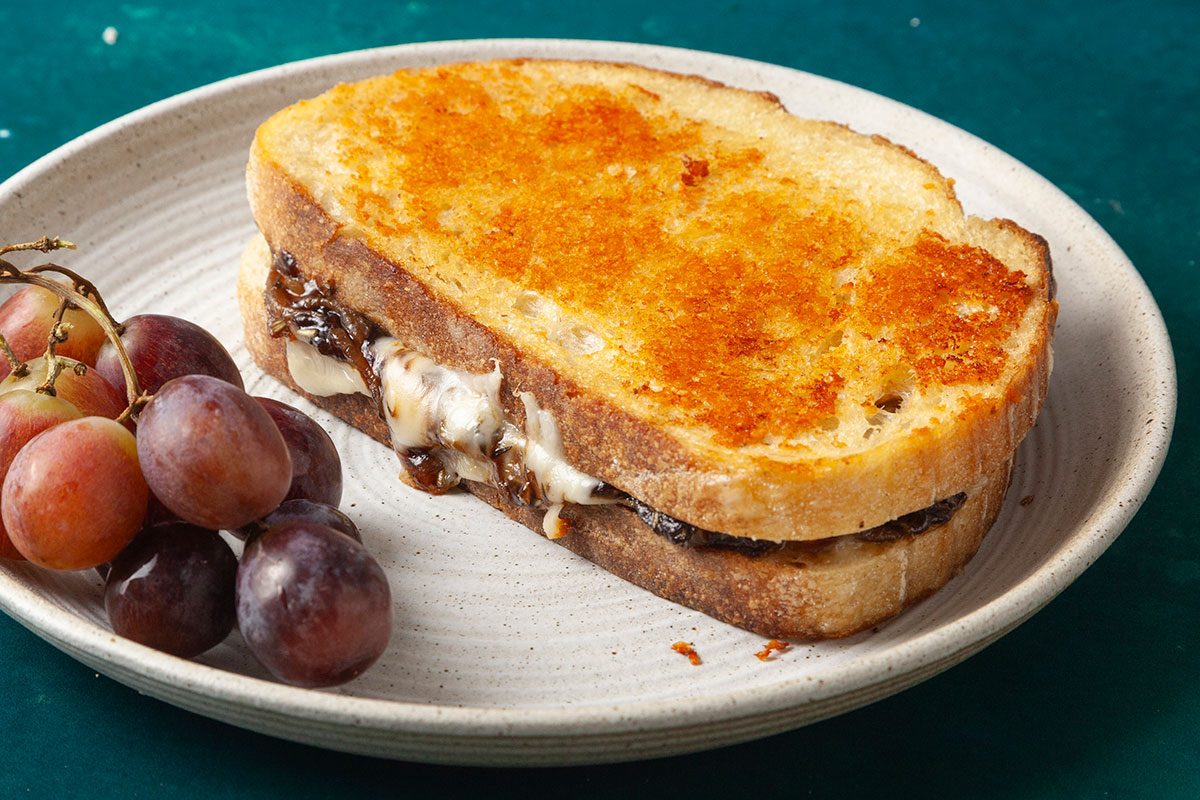 3/4 angle view shot of a French Onion Grilled Cheese sandwich served on a light ceramic plate; The thick slices of bread are toasted to a deep golden brown, with caramelized onions and melted cheese oozing from the center; A small bunch of red grapes sits alongside on a teal surface, completing the cozy, savory presentation;