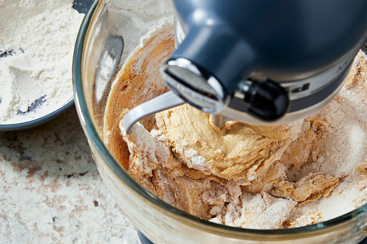 A stand mixer with a dough hook attachment is mixing flour and dough in a glass bowl on a kitchen countertop. A bowl of flour sits nearby.