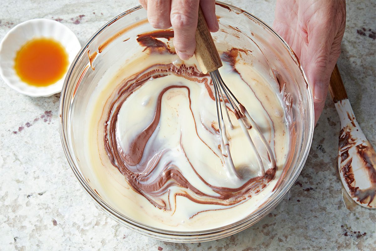 A hand uses a whisk to blend cream and melted chocolate in a glass bowl, creating a marbled pattern. Nearby, a small dish of vanilla extract and a spatula rest on a marble countertop.