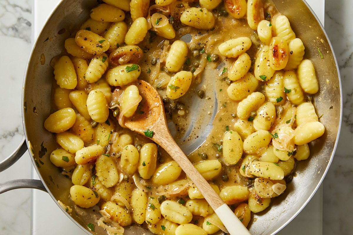 Overhead shot of gnocchi tossed in a buttery caper sauce with herbs, being stirred with a wooden spoon in a skillet;