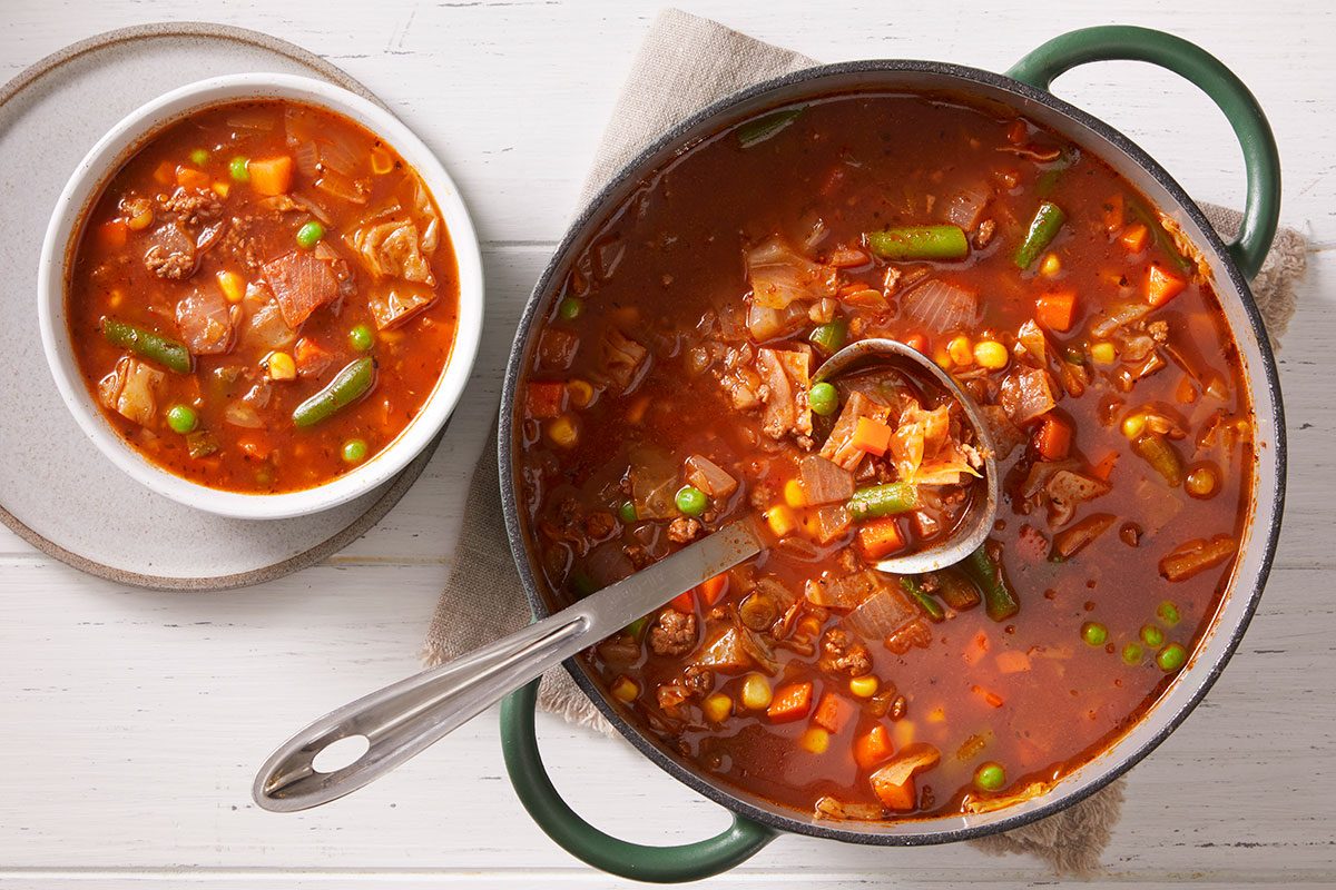 Overhead shot of a pot and a small bowl filled with Italian Veggie Beef Soup soup featuring chunks of beef, green beans, and pasta in a tomato broth; spoon resting nearby.