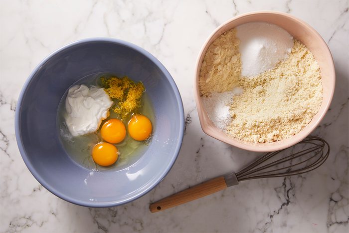 Overhead shot of Two mixing bowls sit on a marble surface one holds eggs lemon zest and yogurt the other has flour sugar and baking powder A whisk is beside them