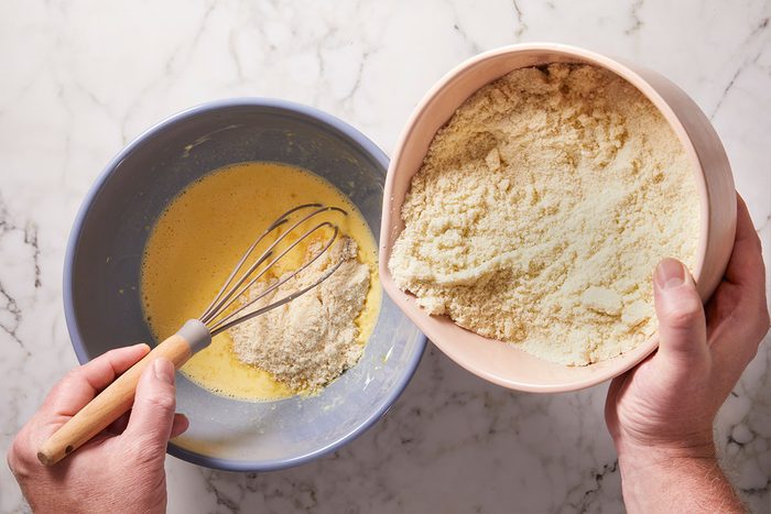 Overhead shot of a person is pouring almond flour from a pink bowl into a blue bowl of whisked eggs on a marble surface as they prepare a mixture