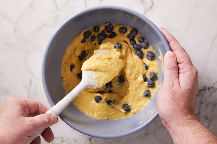 Overhead shot of a person stirs thick batter with fresh blueberries in a light blue bowl using a white spatula on a marble countertop the scene feels calm and inviting