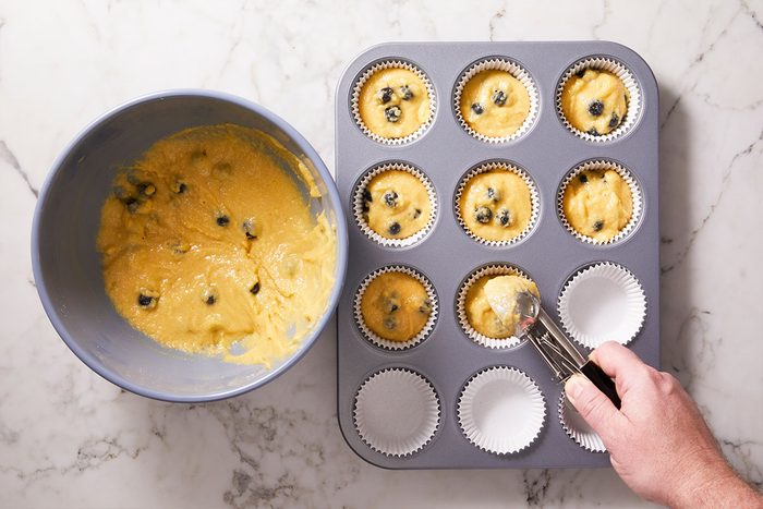 Overhead shot of a hand fills paper cupcake liners with blueberry muffin batter using a scoop and there is a mixing bowl of extra batter on the counter