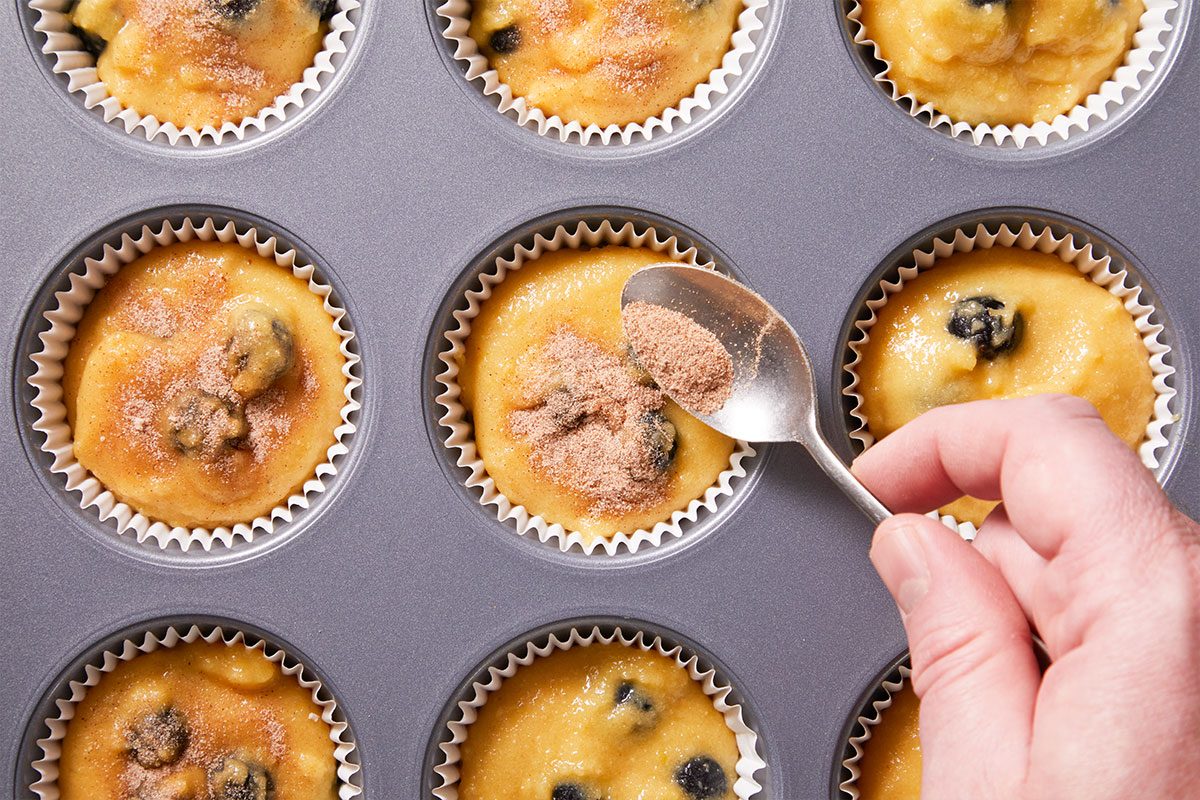 Overhead shot of a hand sprinkles cinnamon sugar over blueberry muffin batter in a metal tin lined with white cups the muffins are not yet baked