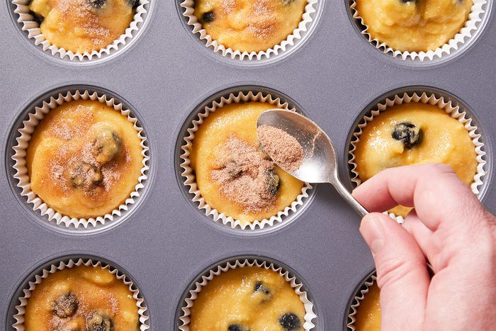 Overhead shot of a hand sprinkles cinnamon sugar over blueberry muffin batter in a metal tin lined with white cups the muffins are not yet baked