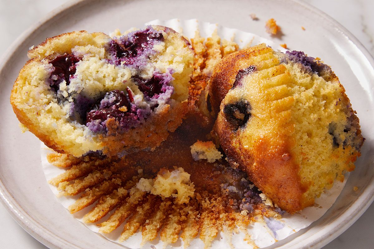 Close-up shot of Keto Blueberry Muffins are shown here sliced open on a white plate with crumbs and a wrapper beneath juicy blueberries appear inside