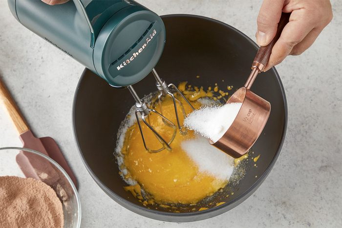 Overhead shot of a hand pouring sugar from a copper measuring cup into a mixing bowl with egg yolks while using a hand mixer, with a bowl of cocoa powder nearby on the countertop;