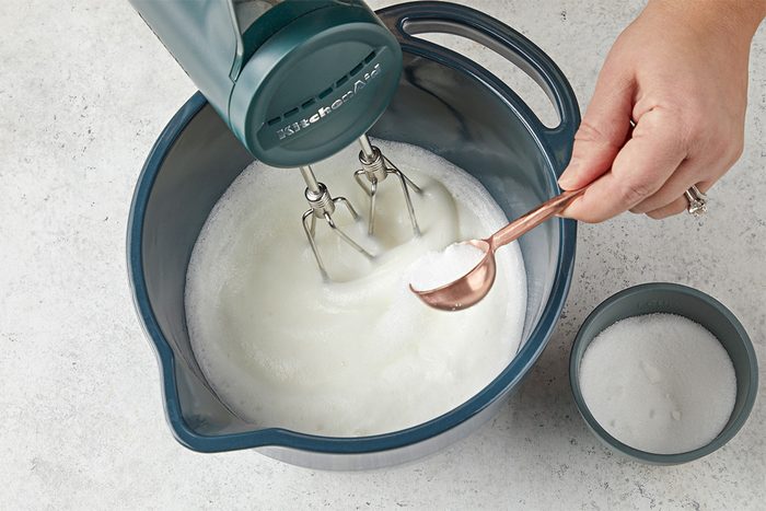 Overhead shot of a person adding sugar from a spoon into a mixing bowl of whipped egg whites while using an electric hand mixer, with a small bowl of sugar nearby on the counter;