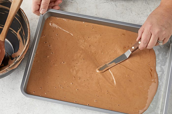 Overhead shot of a person using an offset spatula to spread chocolate cake batter evenly in a rectangular baking pan, with a mixing bowl and wooden spoon nearby;
