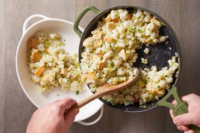 Cooked stuffing mixture being spooned from the skillet into a white baking dish, showing a mix of toasted bread and vegetables.
