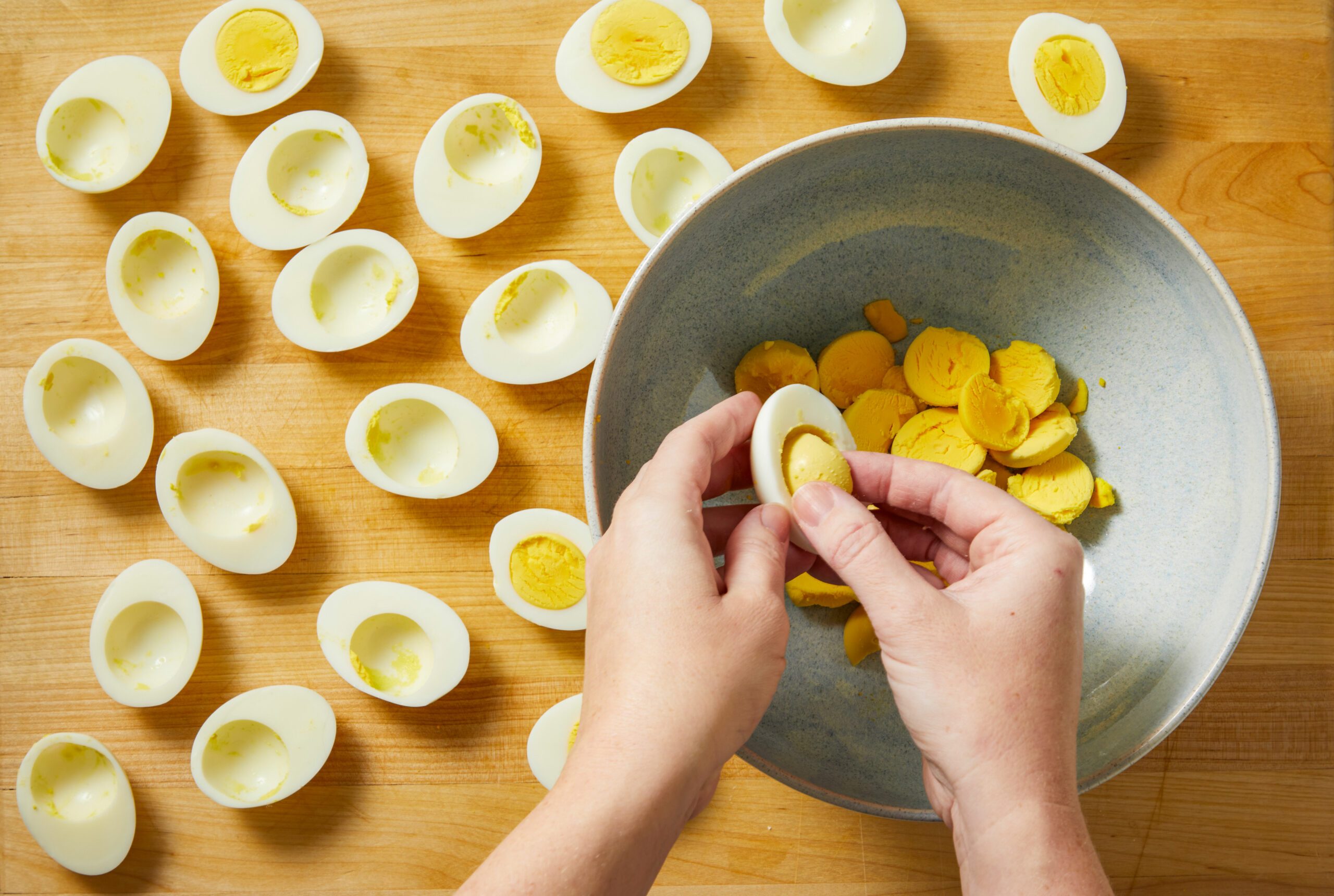 Overhead shot of a person lifts the yolk from a halved hard-boiled egg over a bowl full of yolks More empty egg halves are spread on a wooden surface