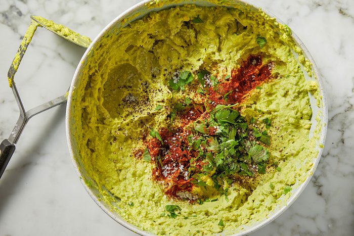 Overhead shot of a bowl of mashed avocado is topped with cilantro black pepper red chili powder and salt It is placed on a marble surface with a potato masher on the side