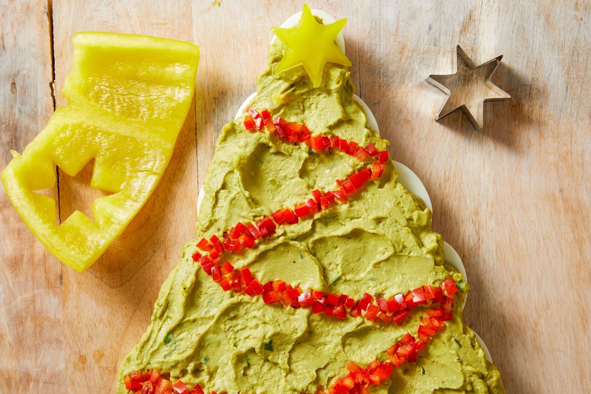 Overhead shot of a platter of guacamole shaped like a Christmas tree is decorated with red and yellow bell pepper A metal cookie cutter sits nearby