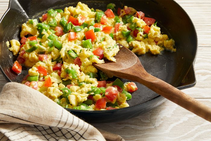 3/4 shot of Vegetable Scrambled Eggs in a black skillet, a wooden spoon resting inside, placed on a striped cloth.