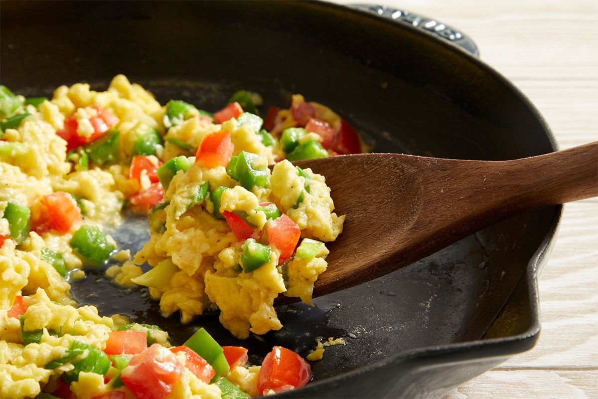 Close up shot of Vegetable Scrambled Eggs in a black skillet, a wooden spoon resting inside, placed on a striped cloth.