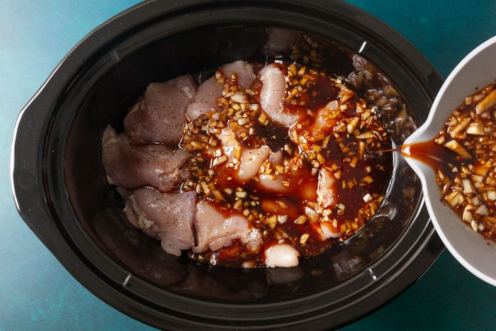 Overhead shot of raw chicken thighs arranged in a slow cooker as a bowl of soy-garlic marinade with diced onions is being poured over the top.