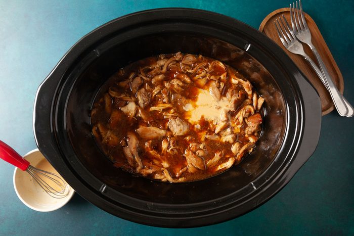 Overhead shot of cooked chicken in a slow cooker simmering in a rich honey-garlic sauce, with a slurry being whisked in a small bowl beside it and a set of forks resting on a wooden tray nearby.