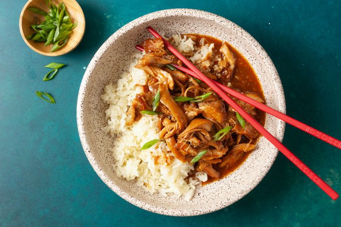 Overhead shot of two speckled bowls filled with white rice topped with tender shredded honey-garlic chicken in a glossy brown sauce, garnished with sliced green onions; chopsticks rest beside the bowls on a teal surface;