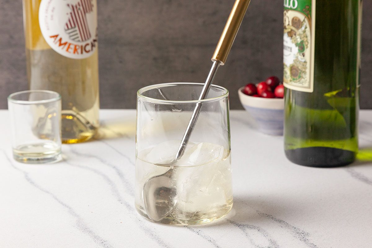 Close-up shot of a clear rocks glass filled with Snowy Negroni cocktail is being stirred, with liquor bottles and fresh cranberries visible in the background.