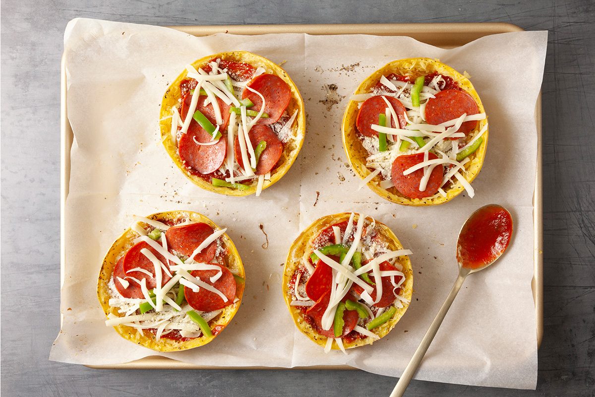 Overhead shot of a parchment-lined baking sheet with four squash pizza halves topped with marinara, pepperoni, shredded cheese, and green pepper strips; A sauce-coated spoon rests beside them on the gray surface;
