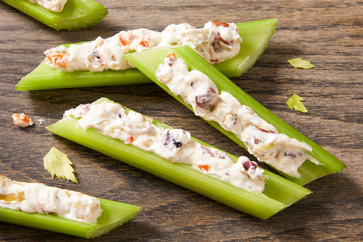 Closeup shot of Olive Stuffed Celery featuring crisp green celery sticks filled with creamy olive-nut spread, arranged on a wooden surface with a few celery leaves scattered around;