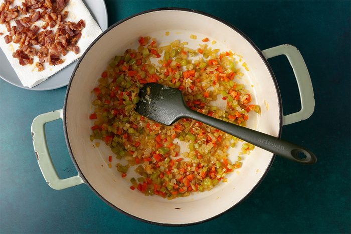 Overhead shot of a pot with sautéed chopped onions, celery, and carrots being stirred with a black spatula, while cooked bacon pieces rest on a paper towel–lined plate beside it;