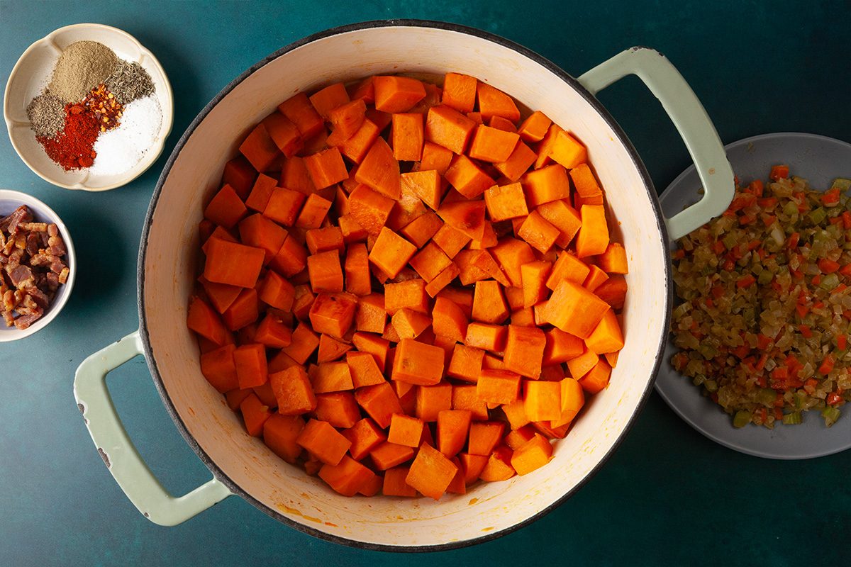 Overhead shot of a large pot filled with cubed sweet potatoes on a green surface, surrounded by small bowls of spices, chopped vegetables, and dried fruit;