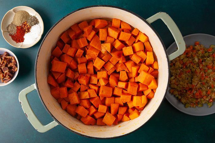 Overhead shot of a large pot filled with cubed sweet potatoes on a green surface, surrounded by small bowls of spices, chopped vegetables, and dried fruit;